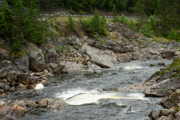 Όμορφο τοπίο με τη νορβηγική βραχώδη βουνό ποτάμι δίπλα στην εθνική οδό με ορμητικά νερά, όπου το νερό σχηματίζει λευκό αφρό. καταρράκτη στο ποτάμι. Πράσινα κωνοφόρα δέντρα στην όχθη του ποταμού. Βροχερή υγρή καλοκαιρινή μέρα. - Φωτογραφία, εικόνα