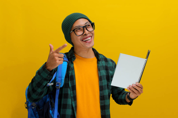 Podekscytowany młody azjatycki student, ubrany w czapkę z kapturem, luźne ubrania, okulary i noszący plecak, wskazuje z entuzjazmem na pustą przestrzeń kopii, stojącą na żółtym tle. - Zdjęcie, obraz