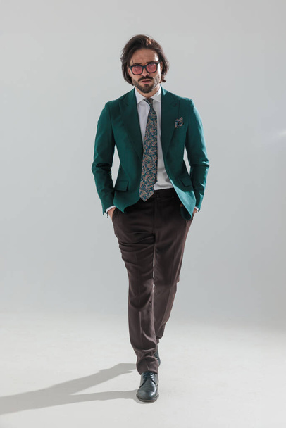 cool uomo d'affari di moda in abito verde con occhiali da sole a piedi con le mani in tasca e guardando avanti davanti a sfondo grigio - Foto, immagini