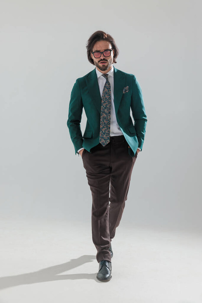pełne ciało zdjęcie eleganckiego biznesmena z okularami przeciwsłonecznymi w zielonym garniturze z brązowymi spodniami spacerujące z rękami w kieszeniach na szarym tle - Zdjęcie, obraz