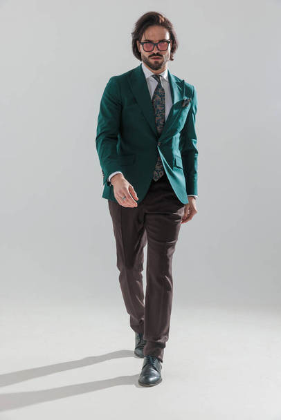 cool fashion man met zonnebril in groen pak lopen in een zelfverzekerde manier in de voorkant van grijze achtergrond - Foto, afbeelding