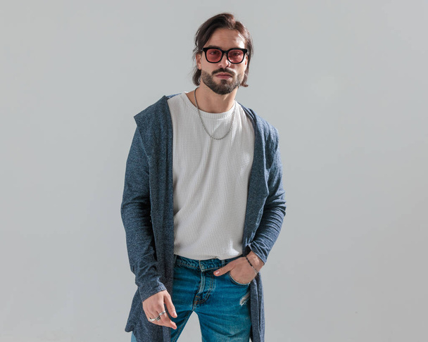 портрет молодого человека в солнцезащитных очках в длинной толстовке, держащегося за руку в синих джинсах и смотрящего вперед на сером фоне - Фото, изображение