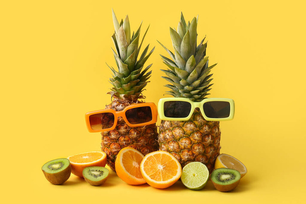 Fresh pineapples with sunglasses, different citrus fruits and kiwi on yellow background - Foto, Bild