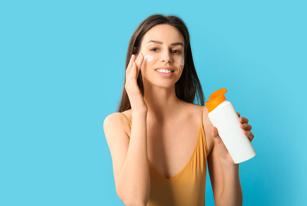 Beautiful young woman applying sunscreen cream on her face against blue background - Foto, immagini