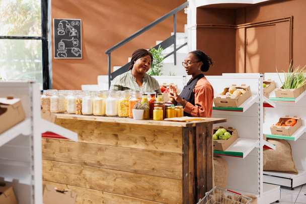 Γυναικείος πωλητής με γυαλιά που παρουσιάζει τα προϊόντα της σε μηδενικά απόβλητα σούπερ μάρκετ, πουλώντας φιλικά προς το περιβάλλον τρόφιμα σε μαύρο καταναλωτή. Τοπικός έμπορος που πωλεί προϊόντα χύμα βιολογικής καλλιέργειας κατά τη διάρκεια της εκδήλωσης - Φωτογραφία, εικόνα
