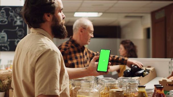 Sıfır atık süpermarketinde yeşil ekran akıllı telefon kullanan bir adam sağlıklı yemek tarifi için malzemeleri kontrol ediyor. Yerel bakkaldaki müşteri organik sebze alırken krom anahtar telefon kullanıyor - Fotoğraf, Görsel