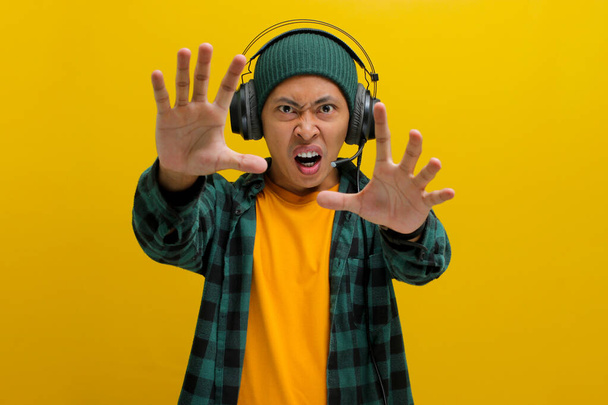 Hombre asiático molesto en un gorro y ropa casual sostiene su mano en un gesto de stop (cámara de mano) mientras escucha música en los auriculares. Aislado sobre un fondo amarillo. - Foto, imagen