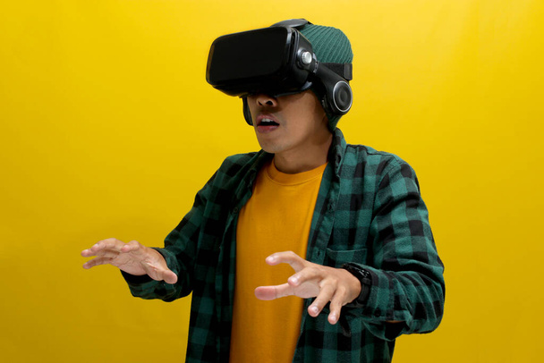 Vzrušený Asiat ve VR sluchátkách natáhne ruku a zdánlivě komunikuje s virtuálním objektem ve VR hře. Izolováno na žlutém pozadí - Fotografie, Obrázek