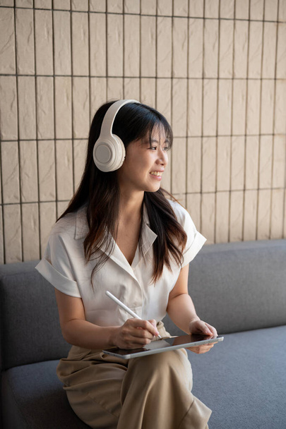 Atraktivní, bezstarostná Asiatka, která nosí sluchátka, ráda poslouchá hudbu a při práci na digitálním tabletu v kavárně se dívá ven. životní styl a technologické koncepty - Fotografie, Obrázek