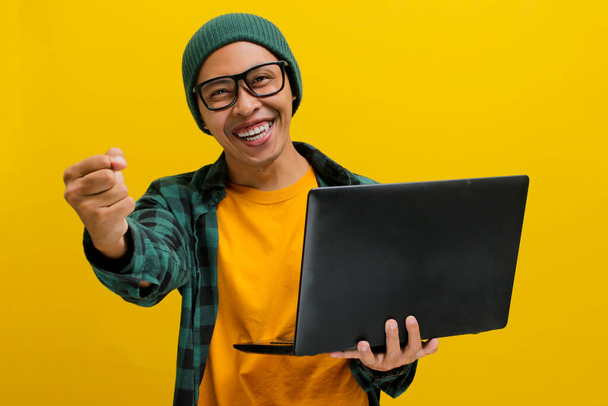 Szczęśliwy młody Azjata, ubrany w czapkę z kapturem i luźną koszulę, podnosi pięść z podniecenia trzymając laptopa, wyrażając gest TAK, świętując rozwój kariery i sukces w biznesie. - Zdjęcie, obraz