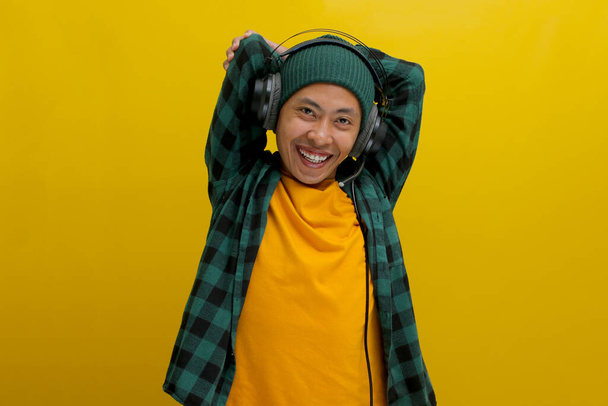 Ασιάτης με σκούφο και καθημερινά ρούχα τεντώνει τους μυς του μετά τη δουλειά, ακούγοντας μουσική στα ακουστικά του. Μια ματιά συγκέντρωσης είναι στο πρόσωπό του. Απομονωμένα σε κίτρινο φόντο. - Φωτογραφία, εικόνα