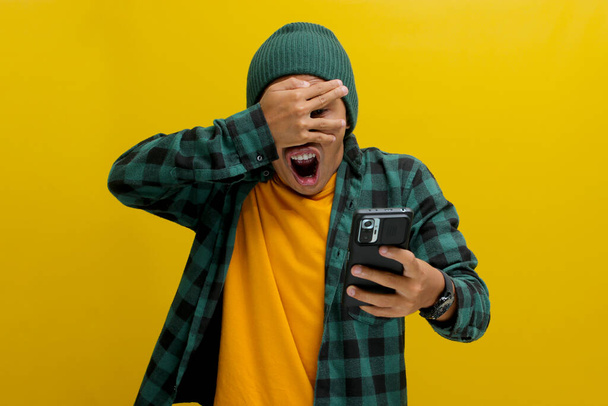 Impresionado hombre asiático, vestido con un sombrero de gorro y camisa casual, cubre sus ojos en una reacción cómica y sorprendida mientras mira su teléfono, claramente sorprendido por noticias inesperadas, fondo amarillo - Foto, Imagen