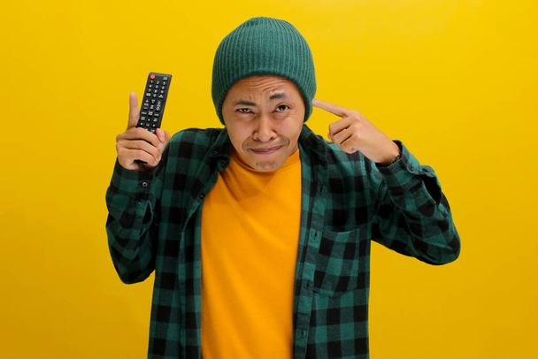Um asiático descontente, vestido com um chapéu de gorro e uma camisa casual, cobre os ouvidos em resposta a ruídos altos enquanto segura um controle remoto da TV, isolado em um fundo amarelo. - Foto, Imagem
