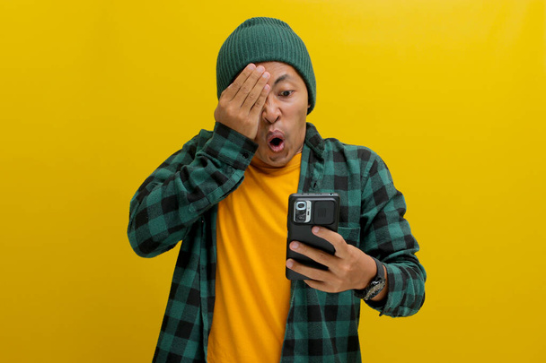 Impresionado hombre asiático, vestido con un sombrero de gorro y camisa casual, cubre sus ojos en una reacción cómica y sorprendida mientras mira su teléfono, claramente sorprendido por noticias inesperadas, fondo amarillo - Foto, Imagen