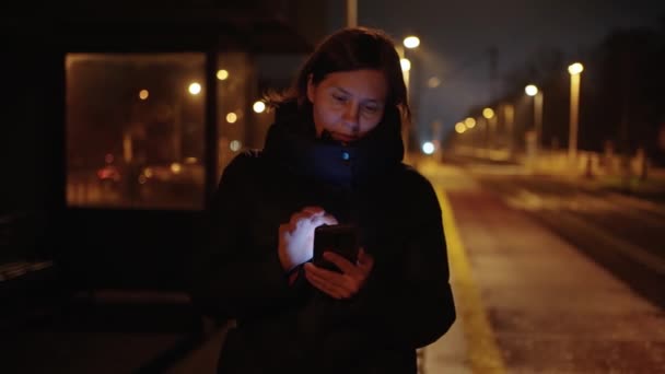 Egy fekete kabátos nő egy városi vonat peronján éjszaka SMS-t használ okostelefonon, miközben a tömegközlekedési megállóban várakozik. - Felvétel, videó