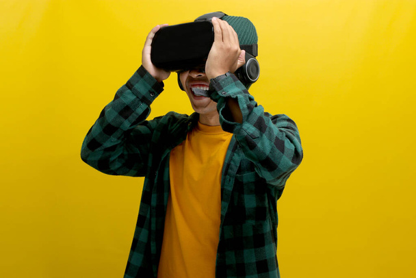 Homme asiatique étonnant portant un casque VR regarde une expérience 3D captivante. Isolé sur fond jaune. - Photo, image