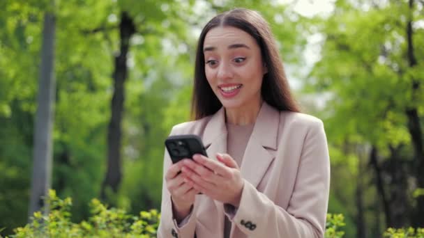 Izgatott nő nézi képernyőn a mobiltelefon a kezében, és így győzelem gesztus, miközben áll a szabadban. A kaukázusi nők örvendeznek, miközben értesítést kapnak az online banki alkalmazástól. - Felvétel, videó