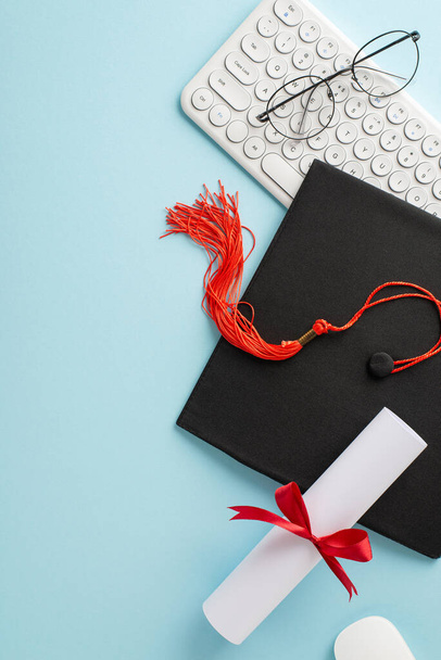 Visão vertical aérea de um boné de graduação, diploma com fita vermelha, óculos e um teclado branco em um fundo azul representando conquistas da educação on-line - Foto, Imagem