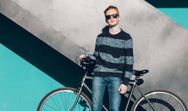Young redhead man standing next to a vintage bicycle in sunglasses, sweater, jeance near turquoise wall warm summer sunny day - Photo, Image