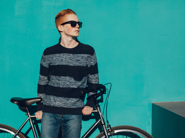 young redhead man standing next to a vintage bicycle in suglasses near turquoise wall warm summer sunny day - Photo, Image
