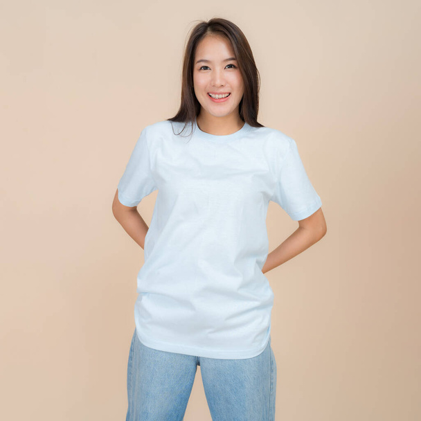 Genç Asyalı bir kadın tarafsız bir bej arka plana karşı duruyor, üzerinde klasik mavi bir kot pantolon bulunan düz bir mavi tişört var, basit ama modaya uygun bir tarzı temsil ediyor. - Fotoğraf, Görsel
