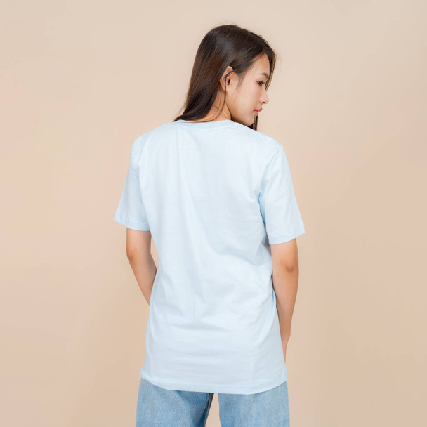 Jonge Aziatische vrouw staat zelfverzekerd tegen een neutrale beige achtergrond, het dragen van een effen blauw t-shirt in combinatie met klassieke blauwe jeans, vertegenwoordigen een eenvoudige maar modieuze stijl - Foto, afbeelding