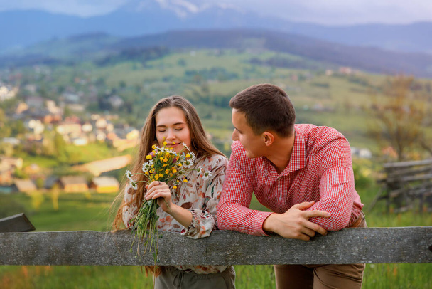 Μια νεαρή γυναίκα και ένας άντρας στέκονται απέναντι σε μια ακτίνα. Πίσω τους είναι ένα όμορφο τοπίο με θέα τα βουνά. Η γυναίκα κρατάει ένα μπουκέτο και εισπνέει το άρωμα των λουλουδιών.. - Φωτογραφία, εικόνα