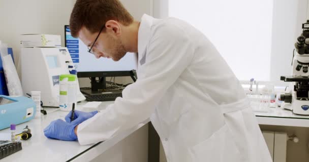 Medical Research Laboratory.Scientist Working Analysing Microbiology Samples.Man in white robe with gloves and glasses putting vial with sample into machine during work in medical laboratory in clinic - Metraje, vídeo