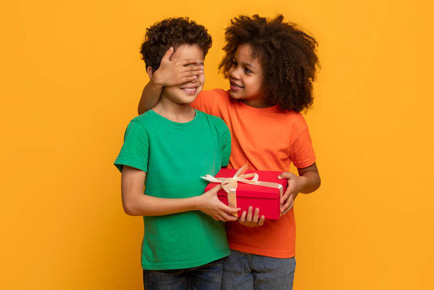 A young African American girl with curly hair is delightfully surprising a boy by covering his eyes with her hand while giving him a red gift box tied with a white ribbon - Photo, Image