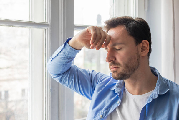 A man standing in front of a window, holding his head with his hands. His facial expression shows signs of distress or deep thought as he looks outside. - Photo, Image