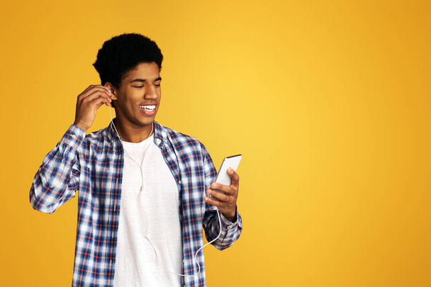 ヘッドフォンと携帯電話、オレンジ色の背景で音楽を聴く幸せなアフリカ系アメリカ人の男 - 写真・画像