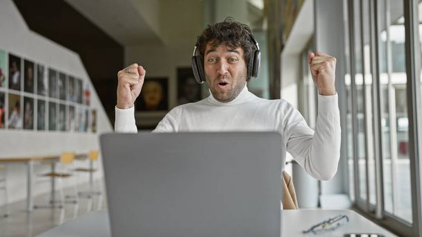 Egy fiatal spanyol férfi az irodában boldogan reagál egy laptop előtt, fejhallgatót visel. - Fotó, kép