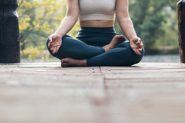 Giovane donna in abbigliamento sportivo meditare e fare yoga in natura. Lei è scalza su una piattaforma di legno vicino a un lago - Foto, immagini