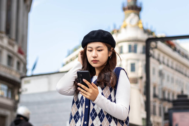 Fiatal felnőtt kínai nő divatos ruhákban okostelefont használ egy európai város utcáin - Fotó, kép