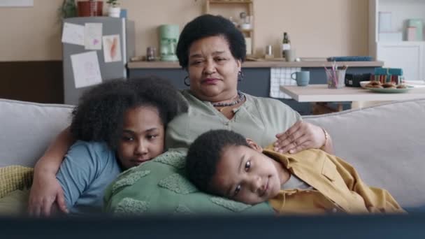 Portré boldog, idős afro-amerikai nőről, aki a kanapén ül, szeretetteljesen ölelgeti nyugodt unokáit, párnákra hajtja a fejét, miközben együtt töltik a hétvégét - Felvétel, videó