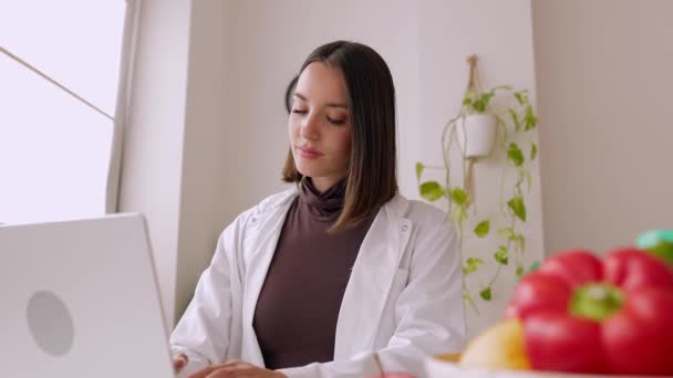Jonge vrouwelijke arts met behulp van laptop zitten op het bureau op medisch consult. Professionele vrouwelijke gezondheidswerker in witte jas aan het werk op de computer op de werkplek. - Video