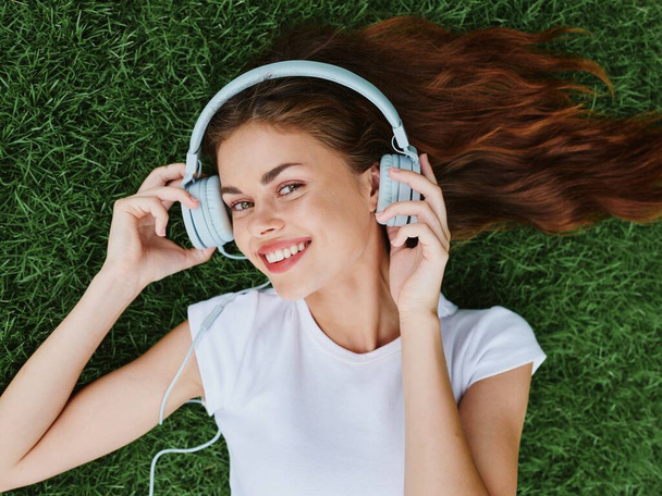 Kobieta nosząca słuchawki leżące na trawie i słuchająca muzyki z uśmiechem na ustach ze szczęściem zębów letnie wakacje w przyrodzie w parku. Wysokiej jakości zdjęcie - Zdjęcie, obraz
