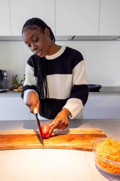 This image captures a young Black woman focused on preparing a healthy meal in a modern kitchen. She is slicing a tomato with care on a wooden chopping board, next to a bowl of freshly grated carrots - Foto, immagini