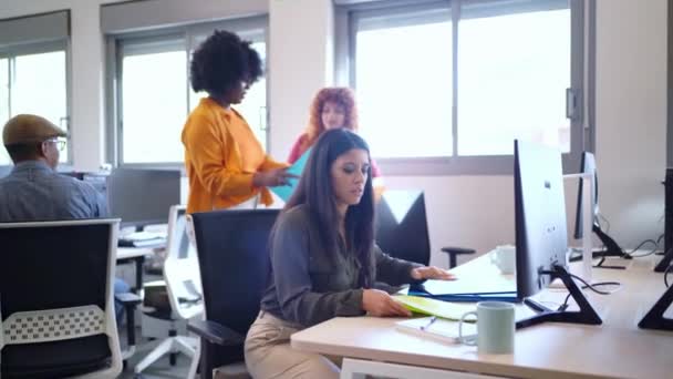 Konzentrierte Frau, die an ihrem Schreibtisch in einem modernen Büro arbeitet. Kollegen interagieren und organisieren Dokumente im Hintergrund.  - Filmmaterial, Video
