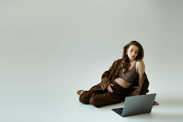 Μια νεαρή έγκυος γυναίκα με καφέ κοστούμι με ένα σακάκι που κάθεται στο πάτωμα ενώ εργάζεται σε ένα φορητό υπολογιστή. - Φωτογραφία, εικόνα