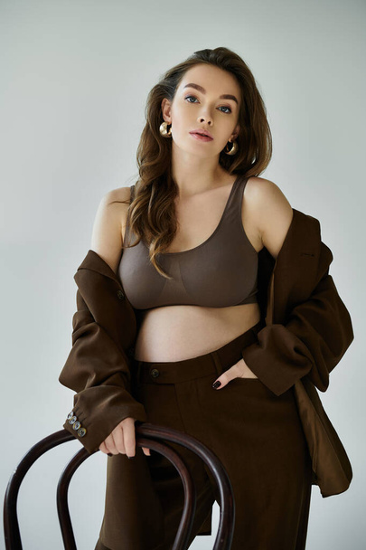 Una joven embarazada exuda gracia en un traje marrón, posando sobre un fondo gris. - Foto, imagen