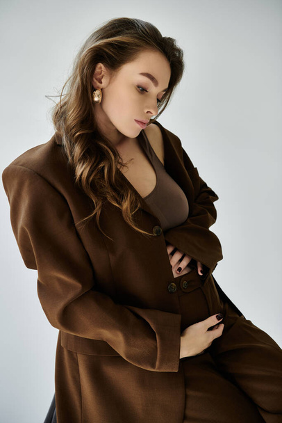 Young pregnant woman in a stylish brown coat poses gracefully for a portrait on a grey background. - Photo, Image