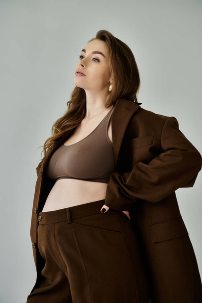 A stylish young pregnant woman in a brown jacket and pants stands gracefully against a grey background. - Fotó, kép