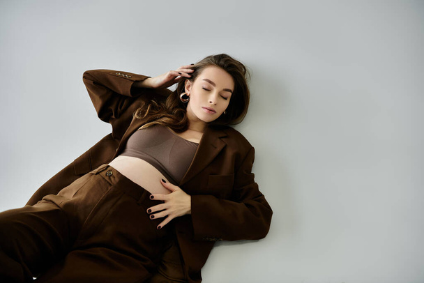 Una joven embarazada con un elegante atuendo marrón, incluyendo una chaqueta, acostada graciosamente sobre un fondo gris. - Foto, Imagen