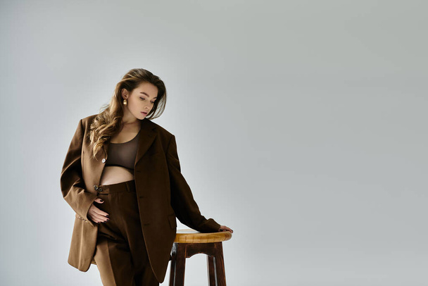 Молодая беременная женщина в коричневом костюме изящно стоит на деревянном стуле на сером фоне. - Фото, изображение