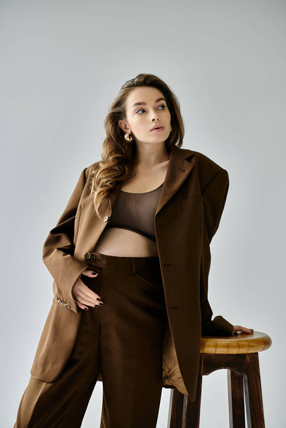 Una joven embarazada con un traje marrón con una chaqueta sentada elegantemente encima de un taburete de madera sobre un fondo gris. - Foto, imagen