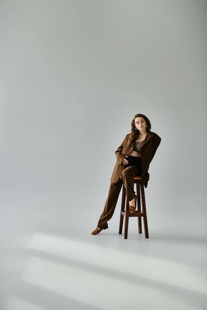 Jeune femme enceinte en costume marron s'assoit gracieusement sur une chaise en bois sur un fond gris. - Photo, image