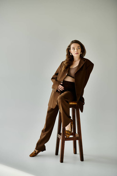 Junge schwangere Frau im braunen Anzug mit Blazer sitzt anmutig auf einem Holzhocker vor grauem Hintergrund. - Foto, Bild