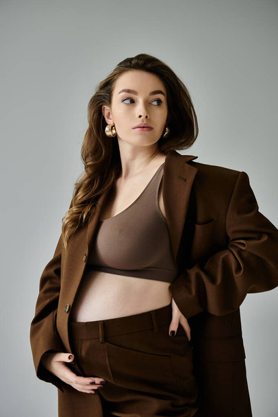 A stylish young pregnant woman in a brown suit striking a pose on a grey background. - Photo, Image