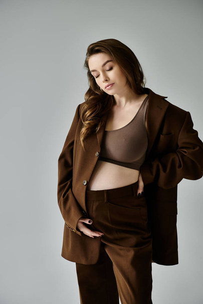 Μια νέα, έγκυος γυναίκα σε ένα κομψό καφέ κοστούμι με ένα σακάκι με αυτοπεποίθηση θέτει σε ένα γκρι φόντο. - Φωτογραφία, εικόνα